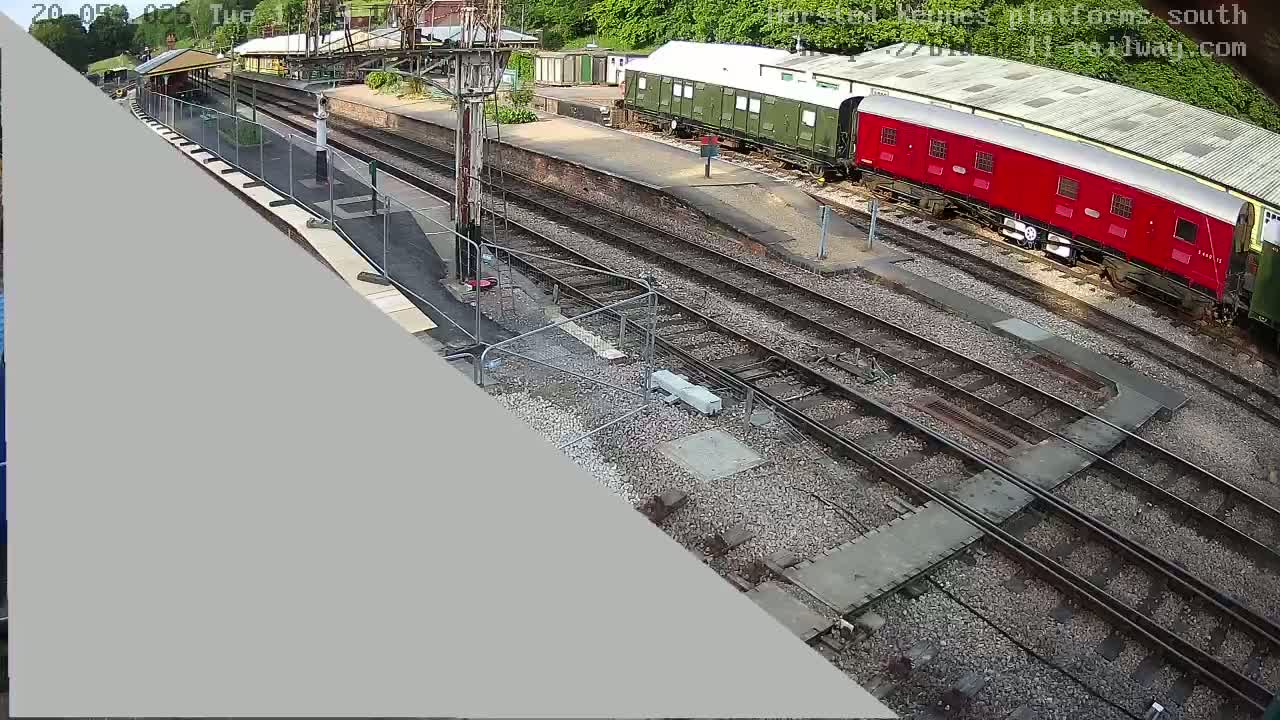 A high-angle view shows a train station with multiple train tracks, a partially obscured red train car, and a green train car under a partly sunny sky.