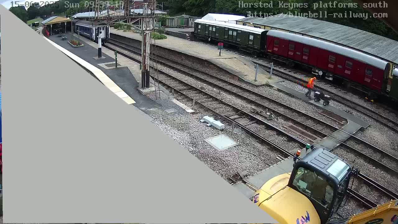 A railway station with several train cars, a worker pushing a cart, and a yellow construction vehicle are visible on a partly sunny day.