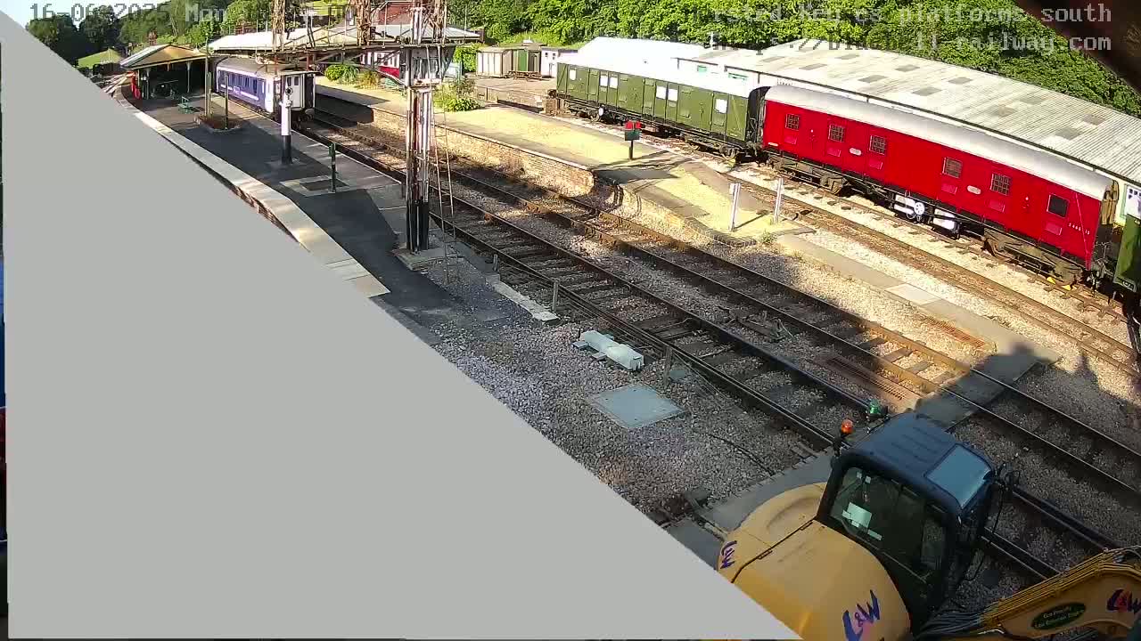 A high-angle view shows a railway station with several train cars parked on the tracks on a sunny day, along with a yellow excavator partially visible in the bottom right corner.