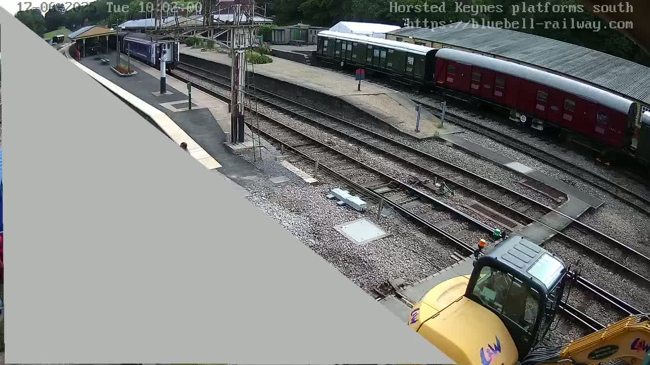 A train station with railway tracks, several train cars, and a yellow excavator is visible under partly cloudy skies.