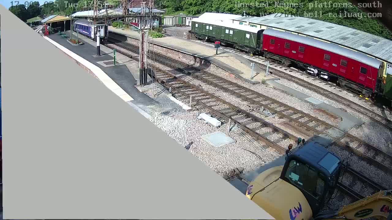 A high-angle view of a railway station on a sunny day shows several train cars, a small building, and a yellow construction vehicle near the tracks.