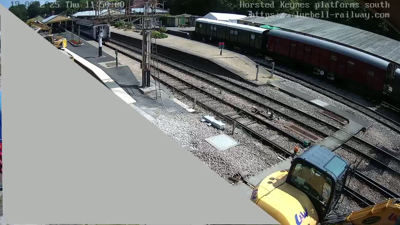 A high-angle view of a train station on a sunny day, showing train cars, tracks, and a yellow construction vehicle near the tracks.