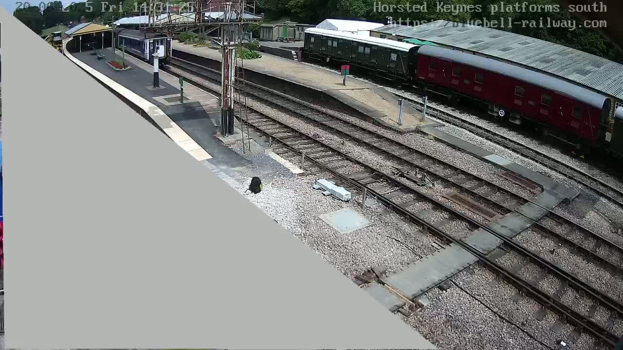 A high-angle view shows a railway station with several train cars, tracks, and a platform on a sunny day.