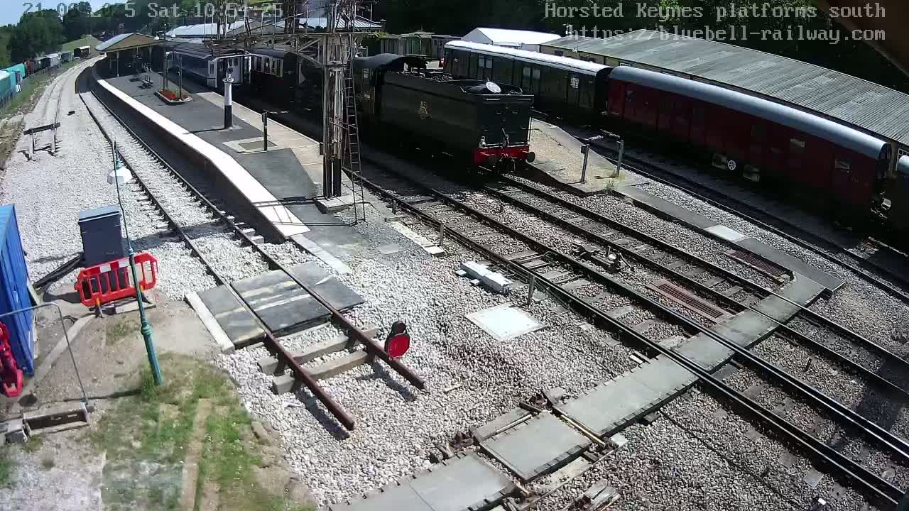A high-angle view of a train station on a sunny day shows a dark-colored steam locomotive and several passenger cars on the tracks, with a platform and other railway infrastructure visible.