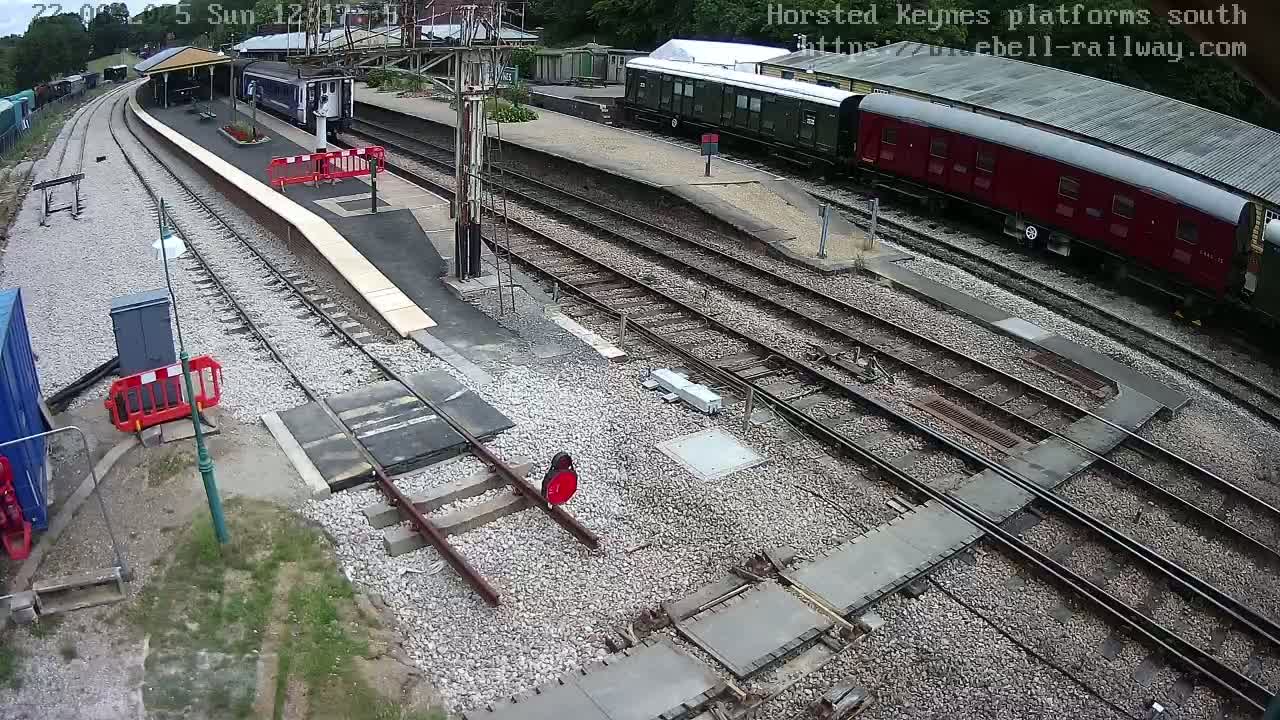An overhead view shows a train station with multiple train cars on several tracks, under a partly cloudy sky.