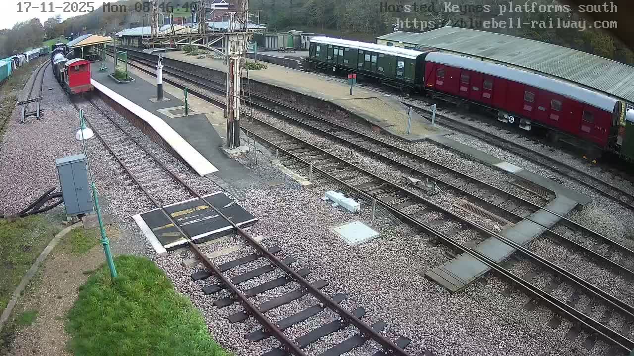 An elevated outdoor view captures a multi-track railway station on an overcast day, with a red locomotive and several passenger carriages parked at platforms, surrounded by gravel ballast and distant trees.