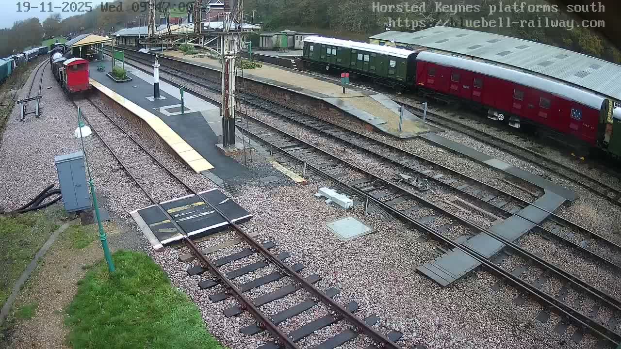 An elevated view of a railway station reveals multiple tracks, a red train car at a platform, and a longer train composed of green and red carriages on a parallel track, all under an overcast sky.