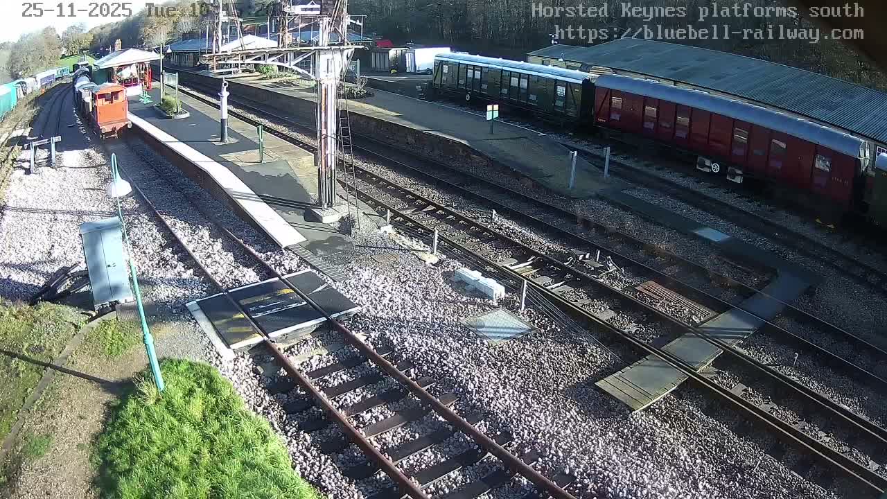 An overhead view captures a bright, sunny day at a multi-track railway station, where an orange shunter locomotive sits at a platform alongside various parked train cars.