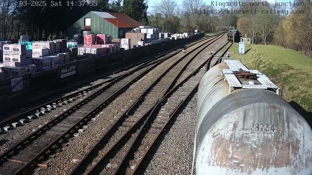 A train passes a railway junction on a sunny day, near a building with stacks of building materials.