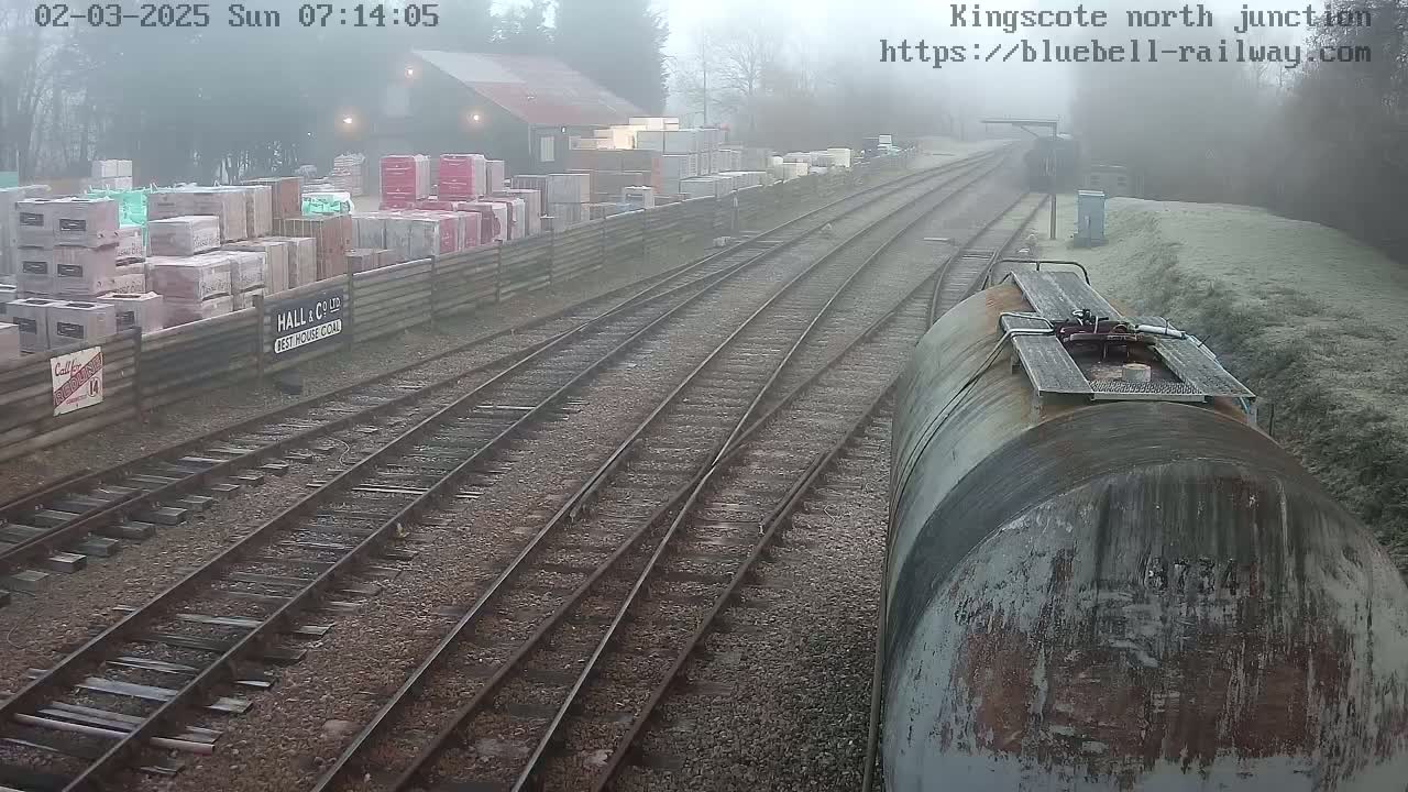 A foggy view of a railway junction with several train tracks, a large cylindrical tank car, and a stack of building materials beside a fence.