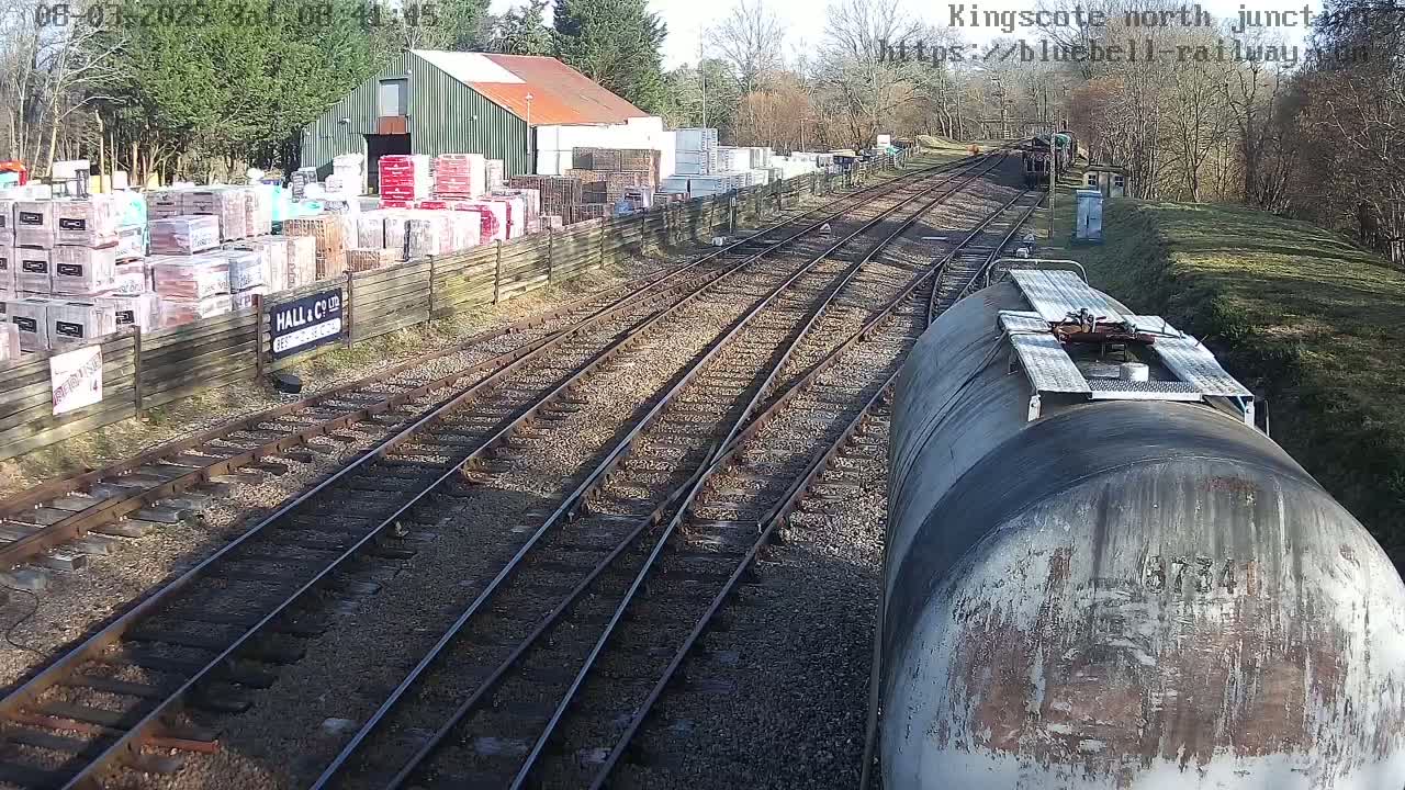 A view from a train shows multiple railway tracks converging at a junction on a sunny day, passing a building with stacked building materials alongside a wooden fence.