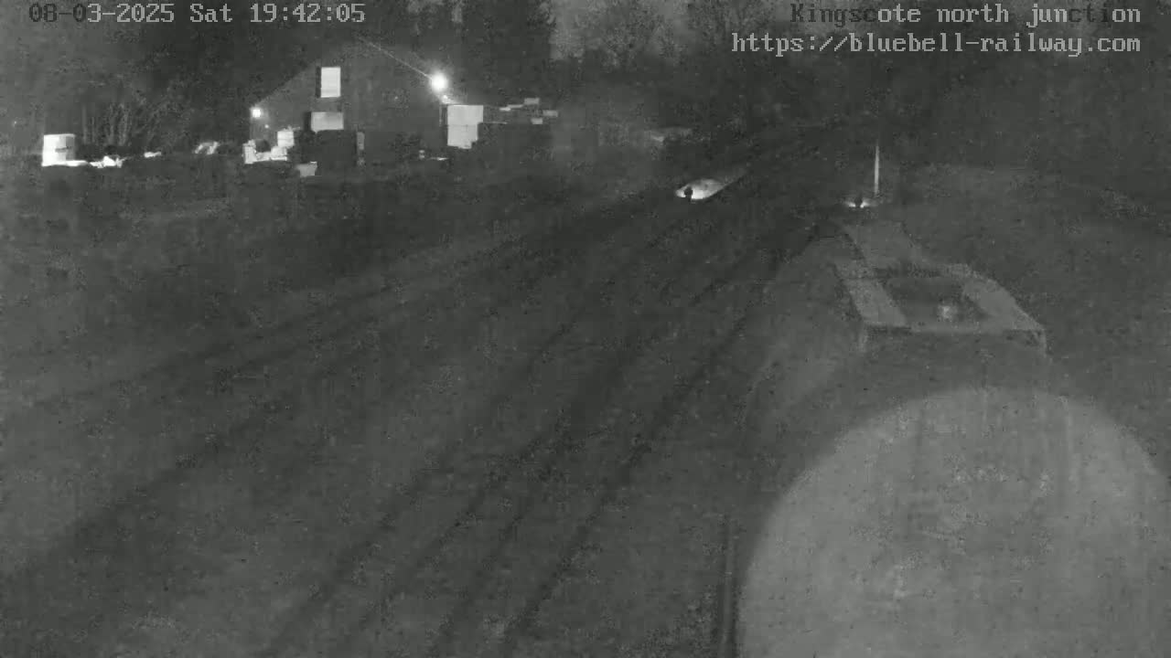 A nighttime, low-light view of railroad tracks and a train partially visible in the foreground, with a building and trees faintly seen in the background.