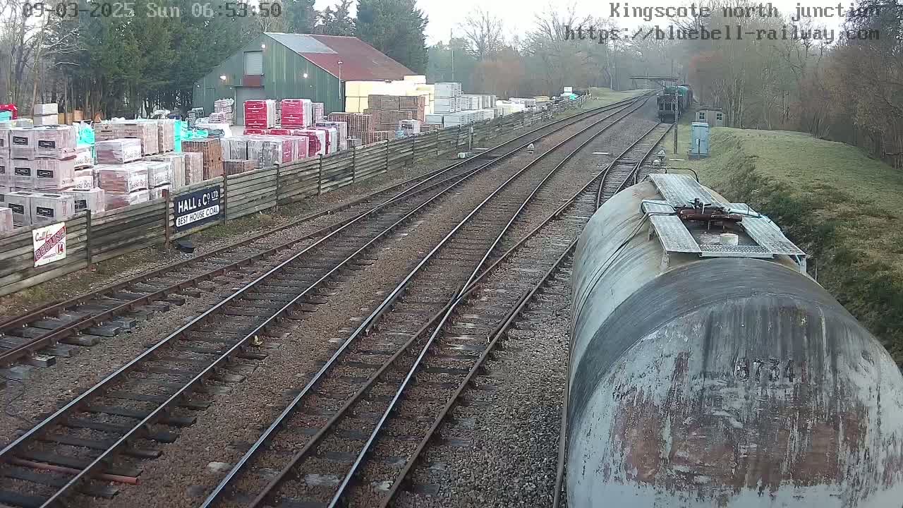 A view from a train car shows several railway tracks converging at a junction, with a building and stacked building materials visible to the side, under an overcast sky.