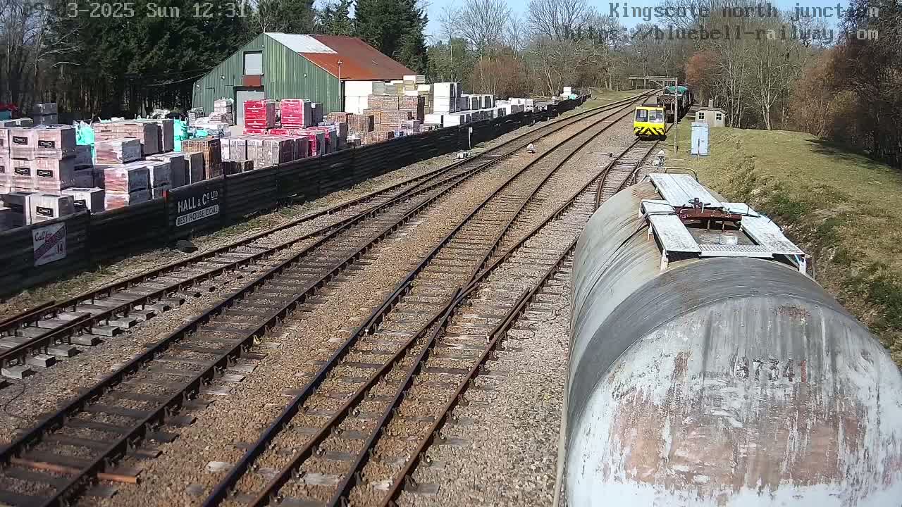 A sunny day shows a railway junction with a small yellow train and a large cylindrical tank car, near a building with stacks of building materials.