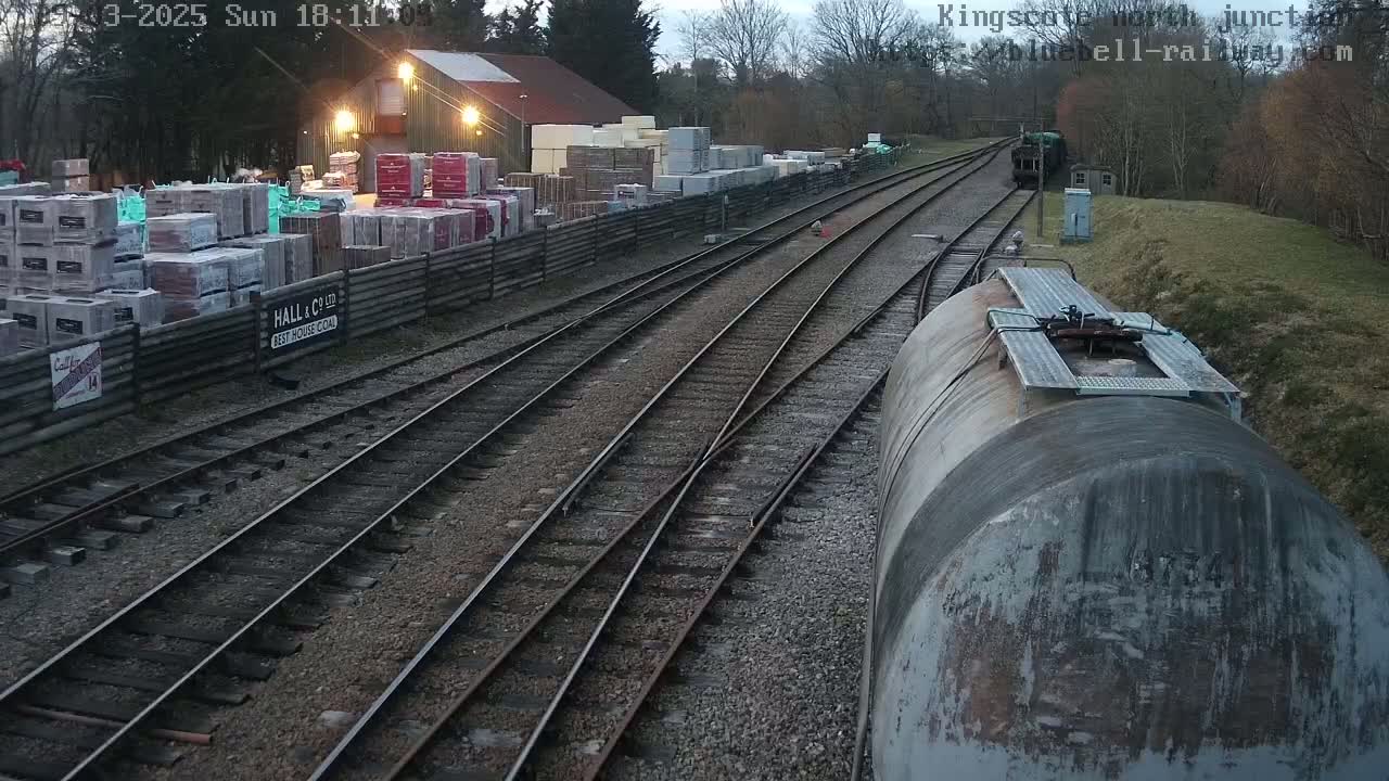 A view from a train shows multiple train tracks leading to a building with stacks of building materials, under a gloomy sky.