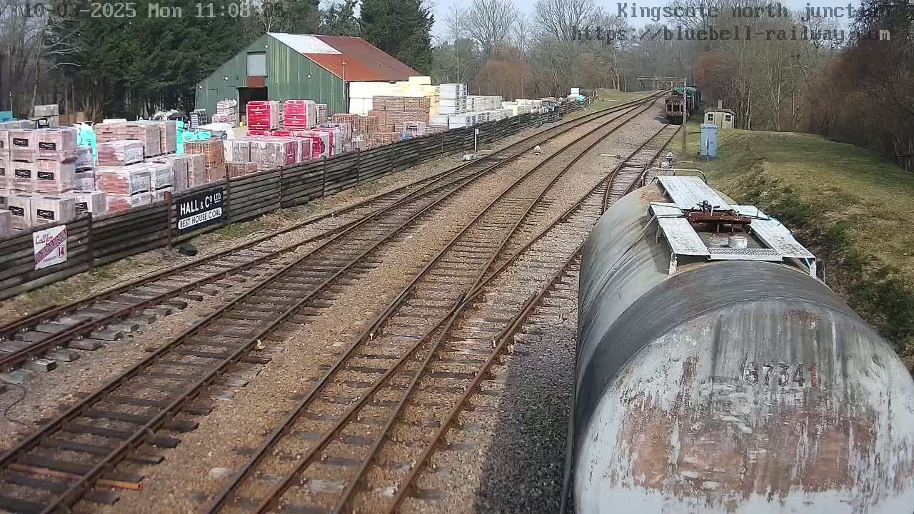 A view from a railway tank car shows a train yard on a partly cloudy day with a building, stacks of bricks, and a wooden fence.