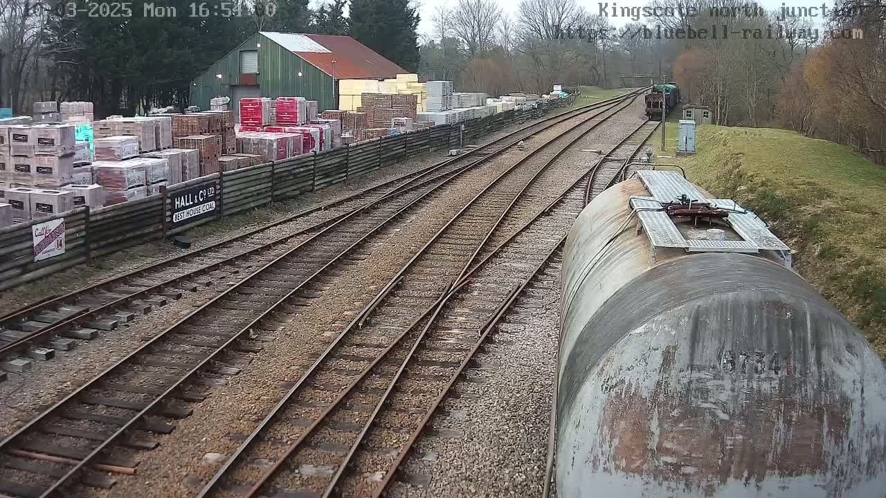 A railway junction on an overcast day, with multiple train tracks converging near a building and stacks of building materials.