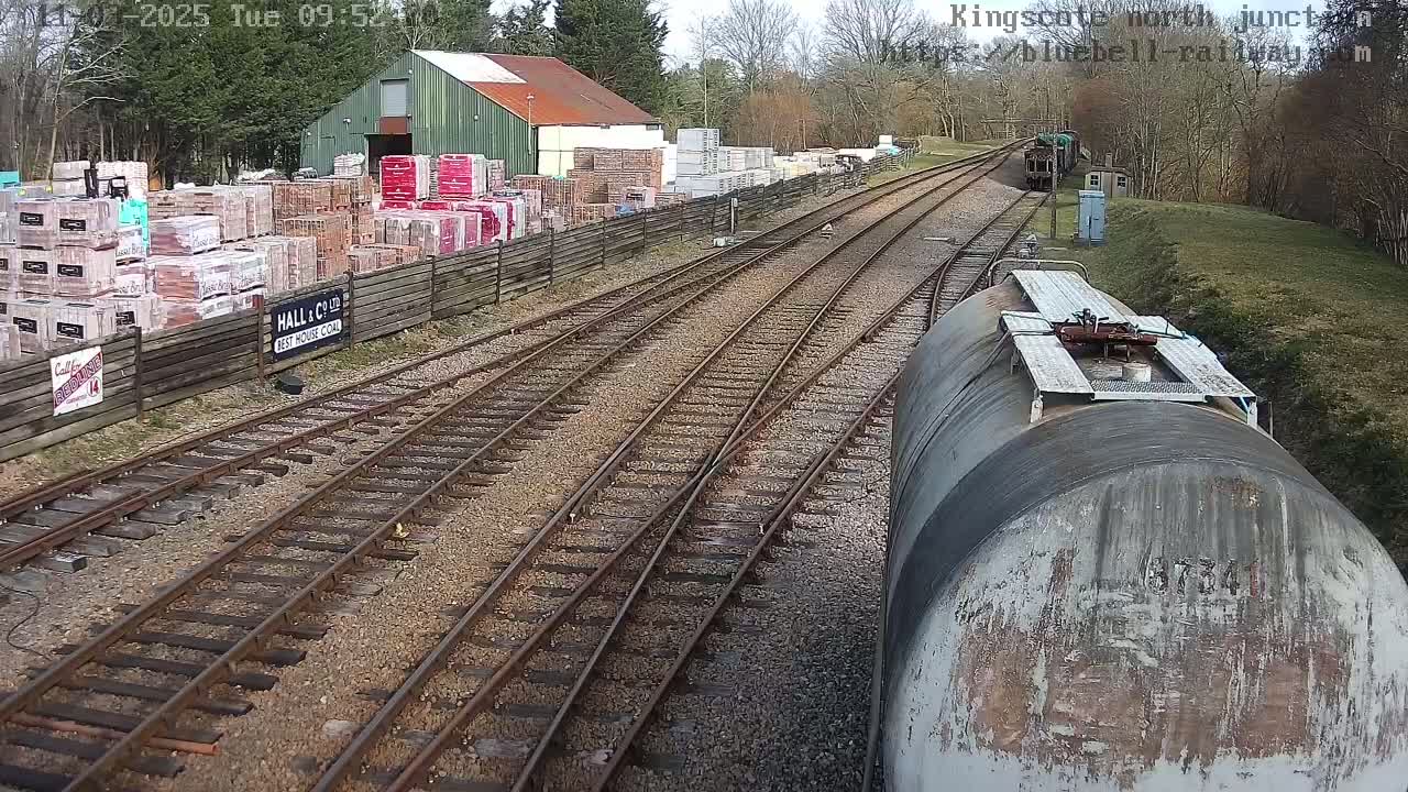 A view from a train car shows railway tracks branching off,  with a building and stacks of bricks visible to one side under a partly cloudy sky.
