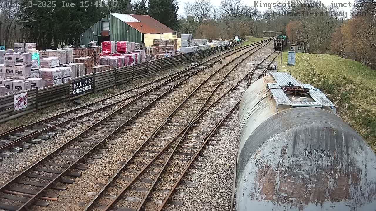 A view from a train shows railway tracks branching off toward a building with stacked building materials, under a partly cloudy sky.