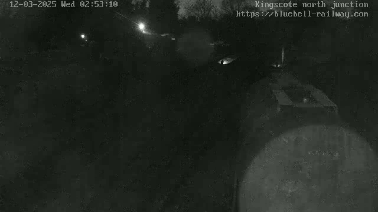 A nighttime, low-light image shows a dark, indistinct area, possibly a train yard or railway junction, with faint lights visible in the distance.