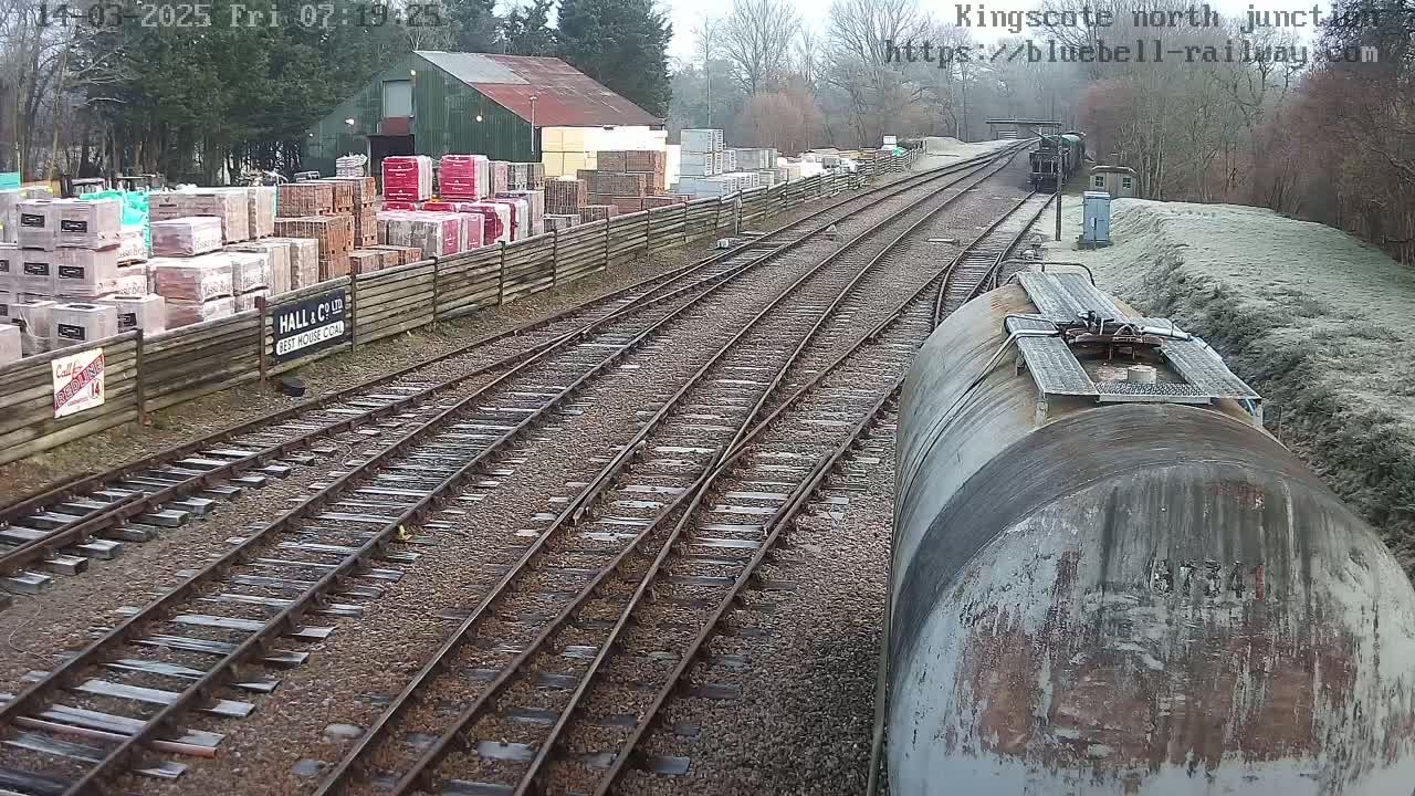A view from a train of railway tracks splitting, with a building and stacks of bricks to the left, and frost on the ground to the right.