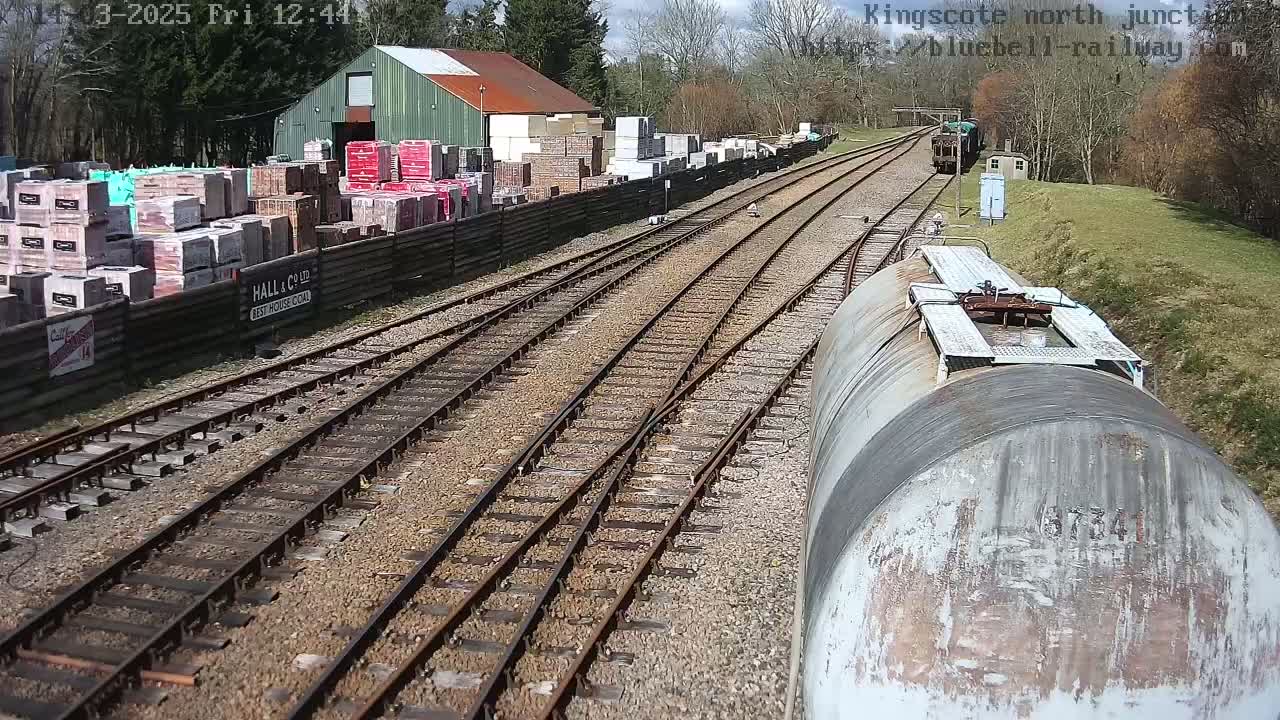 A view from a train of multiple train tracks extending into the distance, past a building and stacks of bricks, on a sunny day.