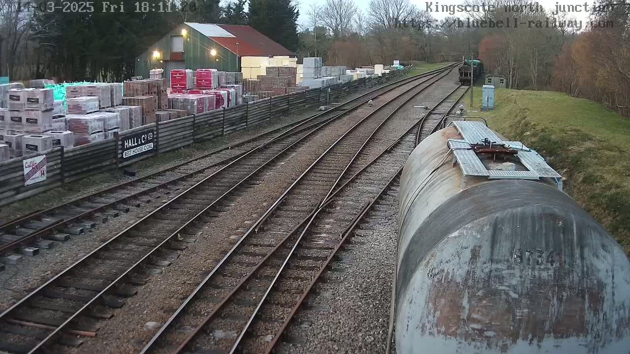 A railway junction viewed from a train, under an overcast sky, shows multiple train tracks splitting off from each other with a building and stacks of building materials visible in the background.