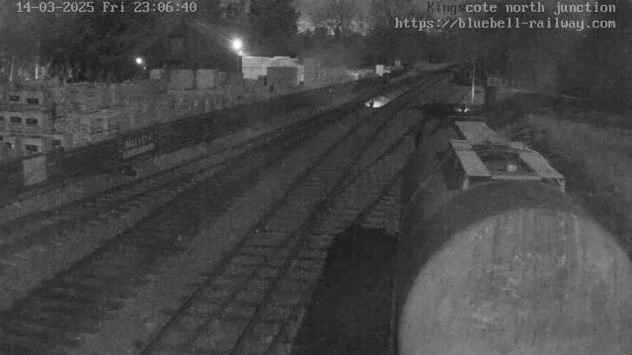 A nighttime view of railroad tracks and a train car, with stacks of materials visible in the background.