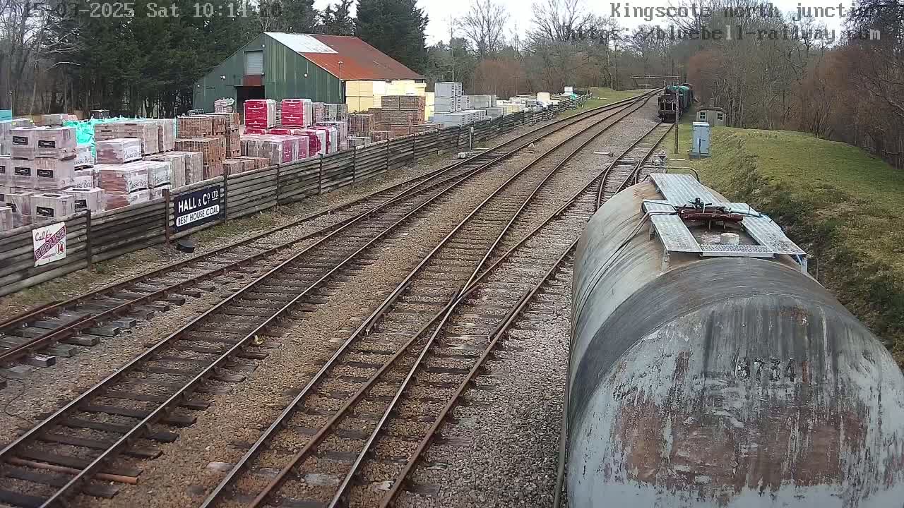 Railway tracks converge at a junction near a building with stacks of building materials, viewed from atop a train car on an overcast day.