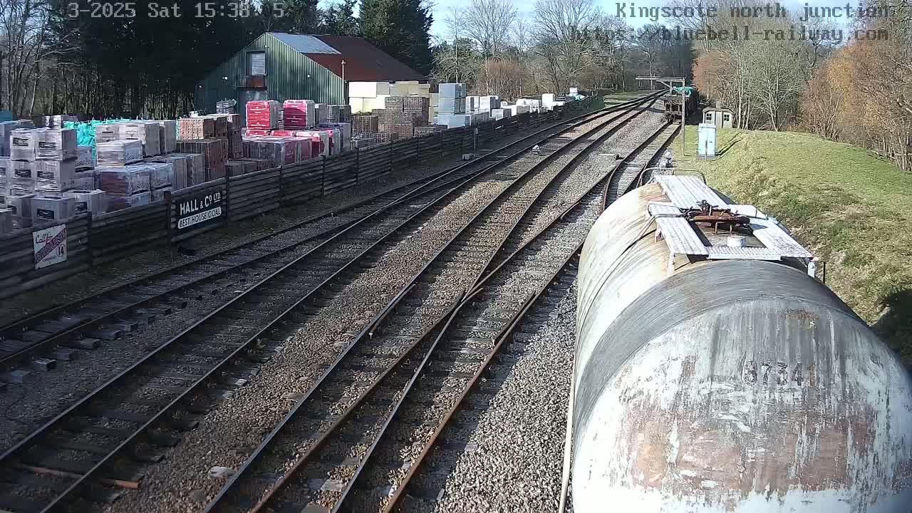A view from a train shows a sunny day at a railway junction with multiple tracks, a building storing stacks of building materials, and a weathered tanker car in the foreground.