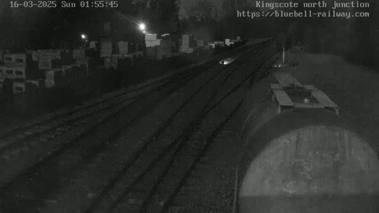 A nighttime view of train tracks, with some structures and stacks of materials visible in the background under dark conditions.