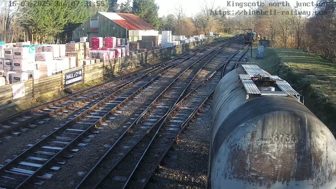 A sunny day at a railway junction shows multiple train tracks converging near a building with stacked materials beside a wooden fence.