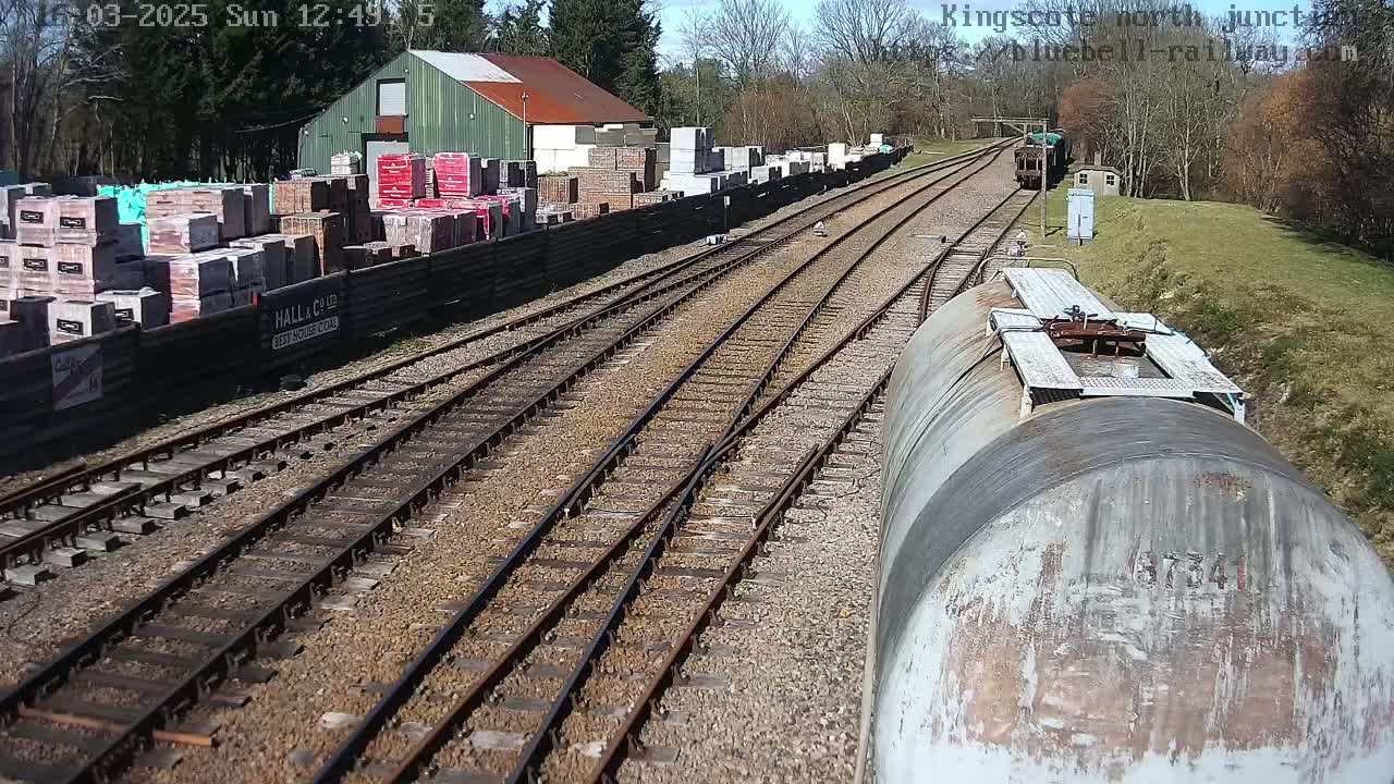 A sunny day shows a railway junction with multiple tracks, a building storing bricks and other materials, and a train car in the foreground.