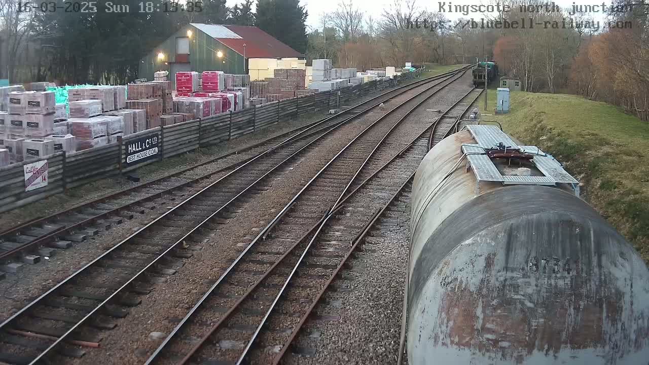 A view from a train car shows railway tracks branching off, passing a building and stacks of bricks behind a fence, under an overcast sky.