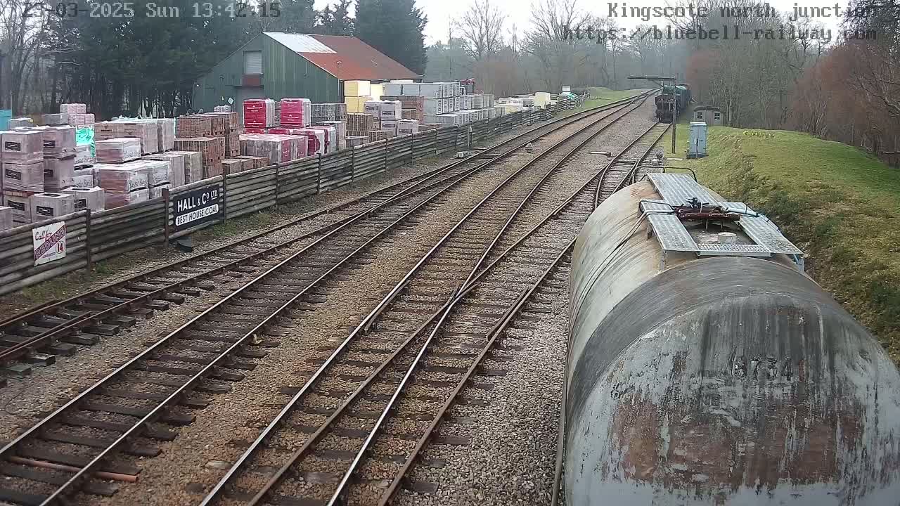 A train sits on a railway track next to a building with stacks of building materials, on an overcast day.