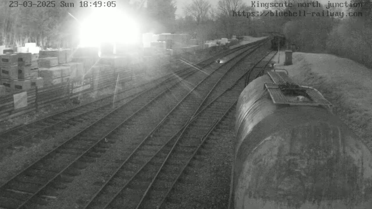 A view from a train of railway tracks diverging from a junction, with a partially visible train car and stacks of materials in the background, under bright, possibly hazy conditions.