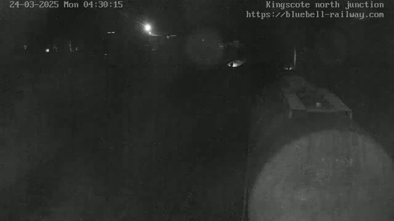 A nighttime, low-light view of a dark area, possibly a railway junction, with faint lights visible in the distance.