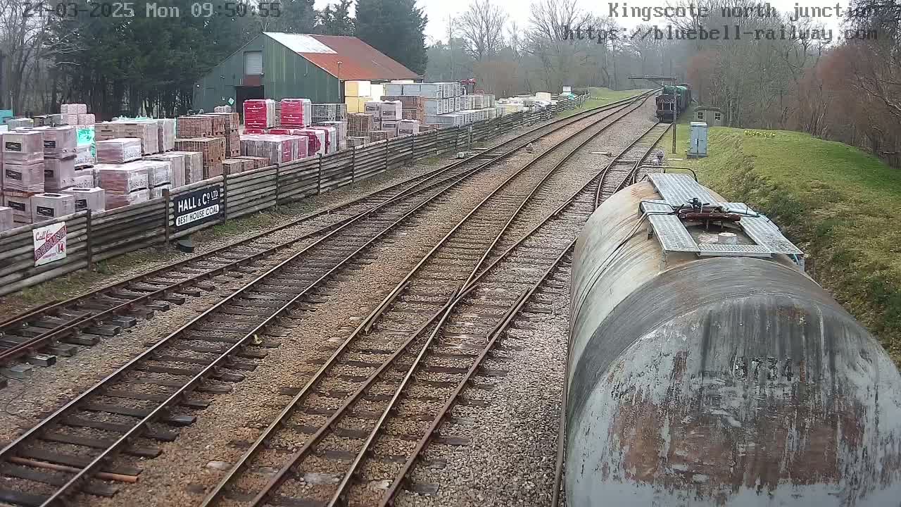 A railway junction with multiple tracks, a train car, and a building with stacks of bricks in the background, under an overcast sky.