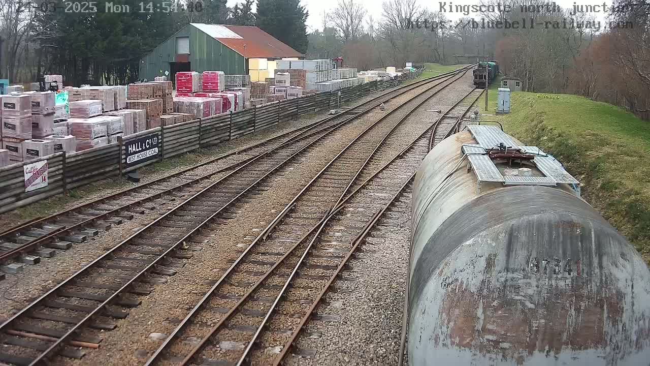 A view from a train shows railway tracks converging, alongside a building with stacks of bricks and a train in the distance, under an overcast sky.