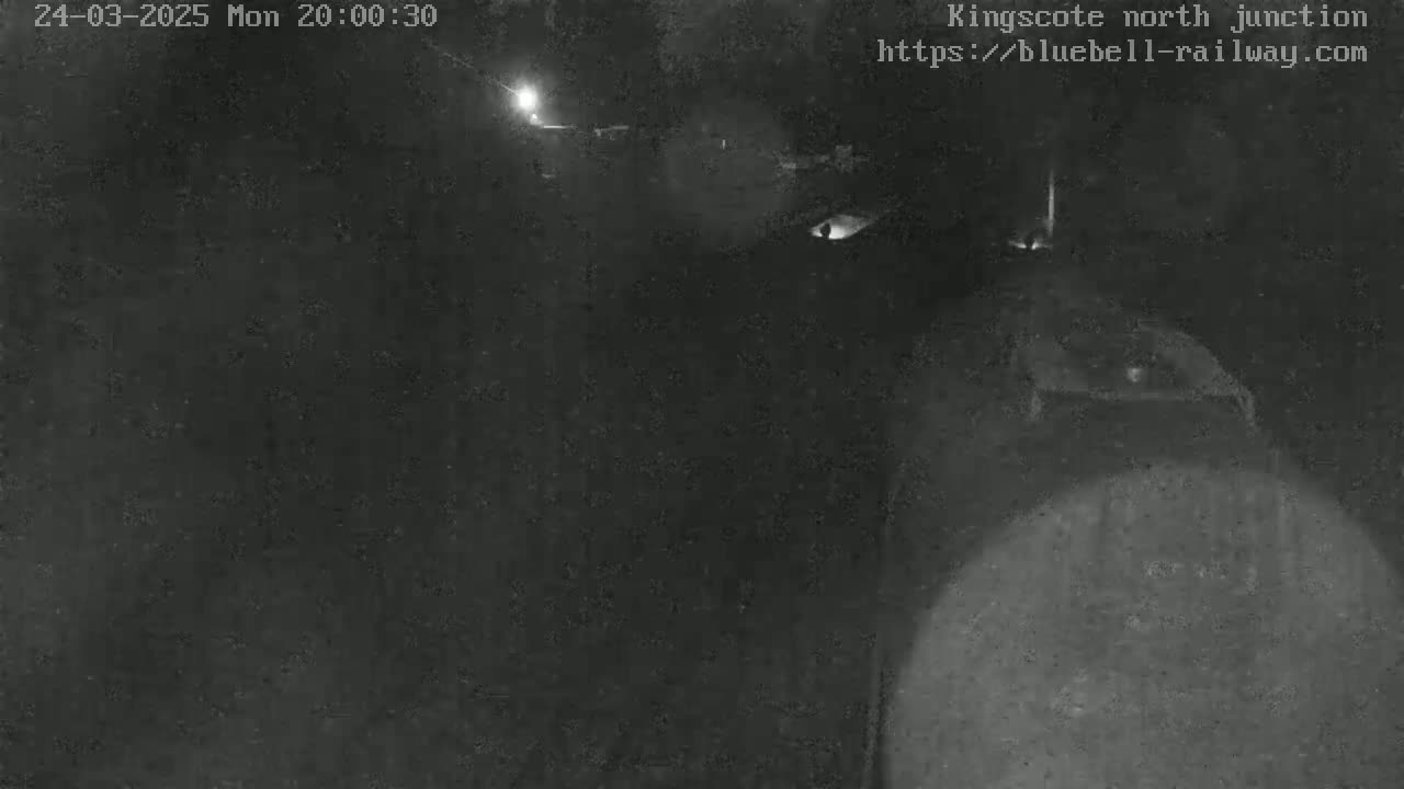 A nighttime, low-light image shows a dark, indistinct area possibly outdoors, with a bright light source in the distance.