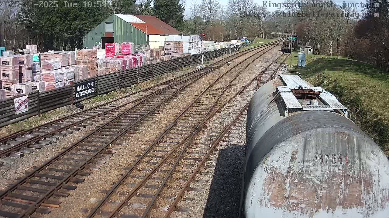 A view from a train car shows railroad tracks splitting into two directions, with a building and stacks of bricks on one side, under a sunny sky.