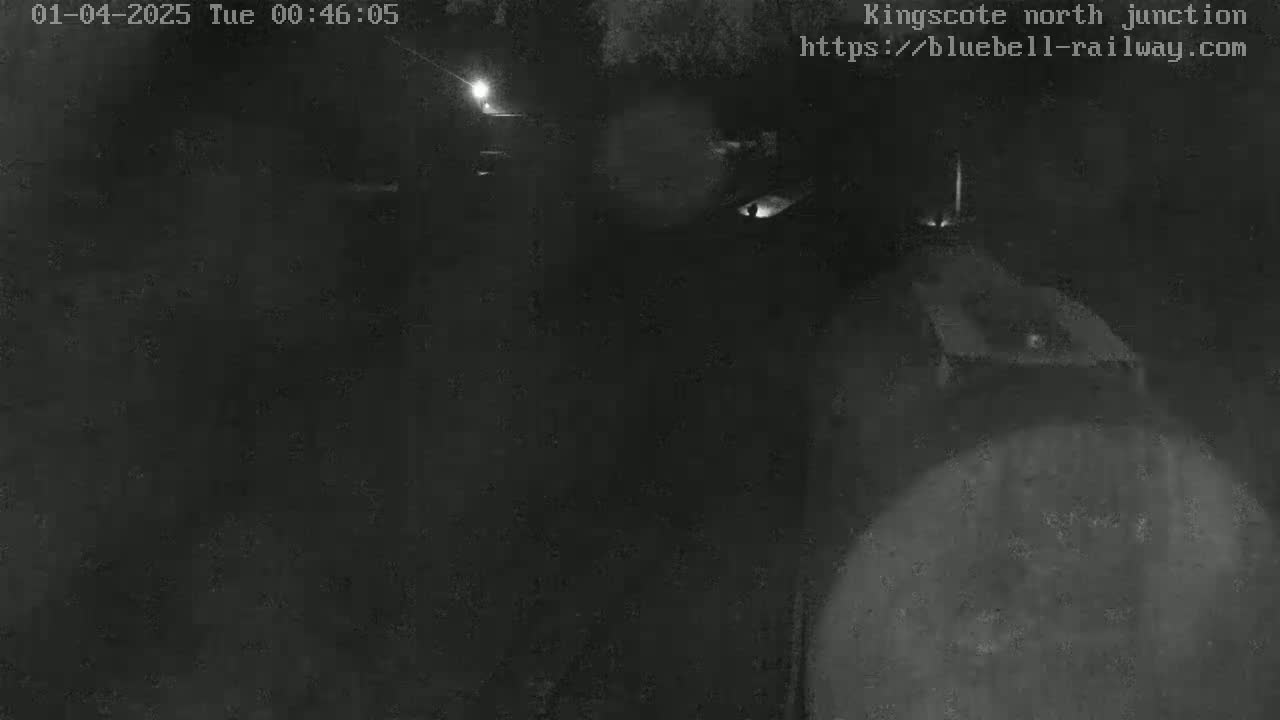 A nighttime, low-light image shows a dark, indistinct area possibly outdoors, with a faint light source in the distance.