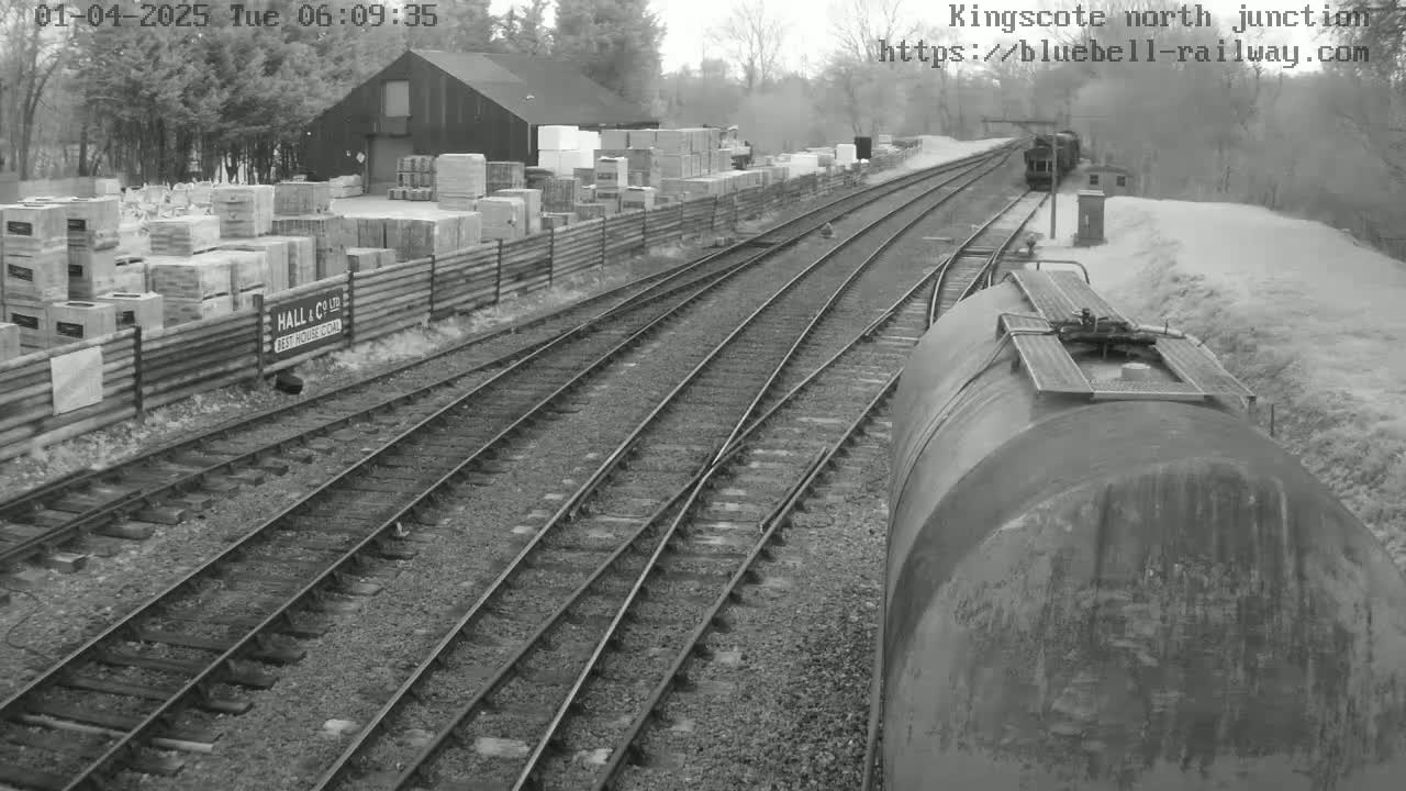 A view from a train shows several railway tracks converging at a junction, with a building and stacked materials alongside the tracks in a snowy landscape.