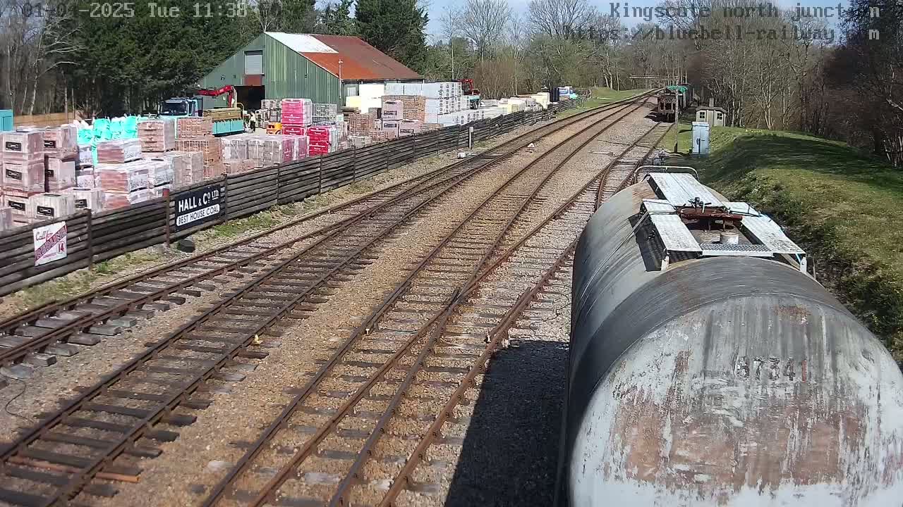 A view from a train shows railway tracks branching off, alongside a building with stacks of bricks and other building materials, under a sunny sky.