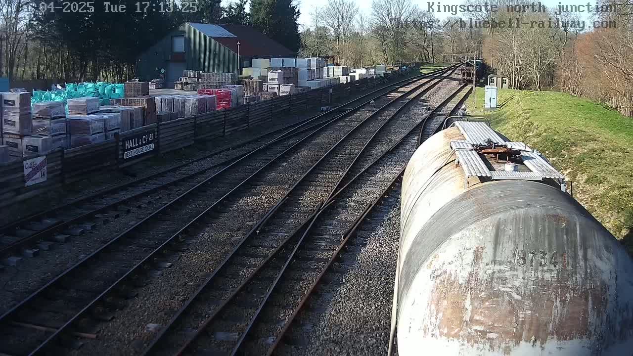 A view from a train car shows railroad tracks splitting in front of a building and a yard filled with stacks of building materials under a sunny sky.
