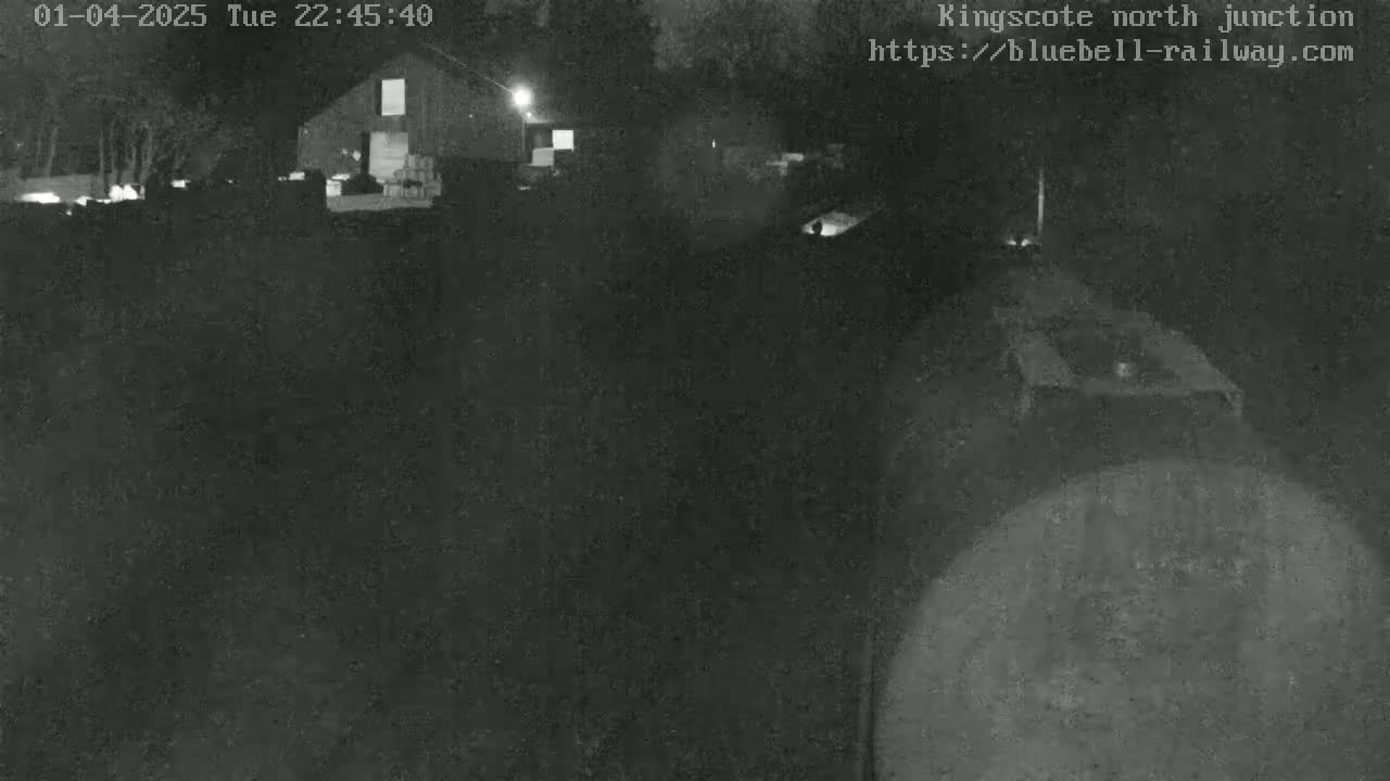 A nighttime, low-light image shows a dark, rural scene with a barn in the background and an indistinct object in the foreground.