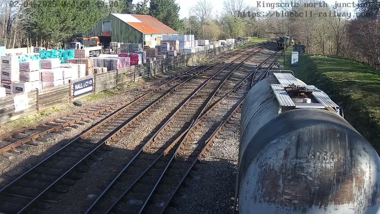 A train passes a railway junction on a sunny day, alongside a building and a yard containing stacked materials.
