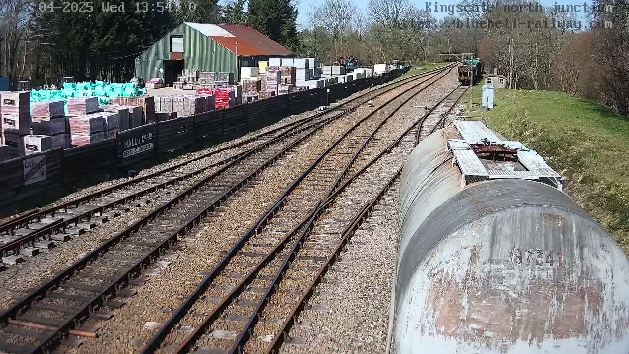A railway junction on a sunny day, with multiple train tracks converging near a building materials yard and a train car in the foreground.