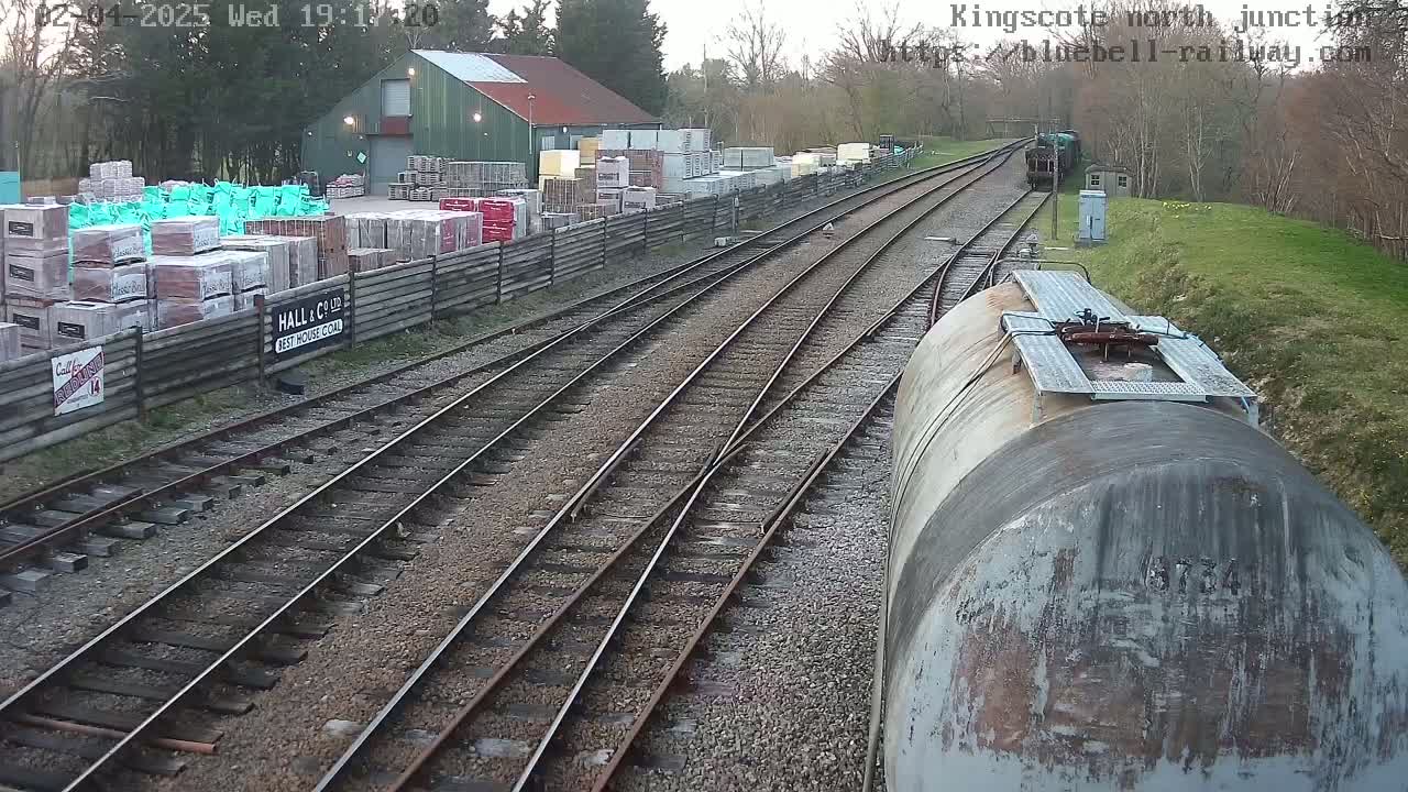 A train car's view of a railway junction, featuring several railway tracks, a building with stacked materials, and a cloudy sky.