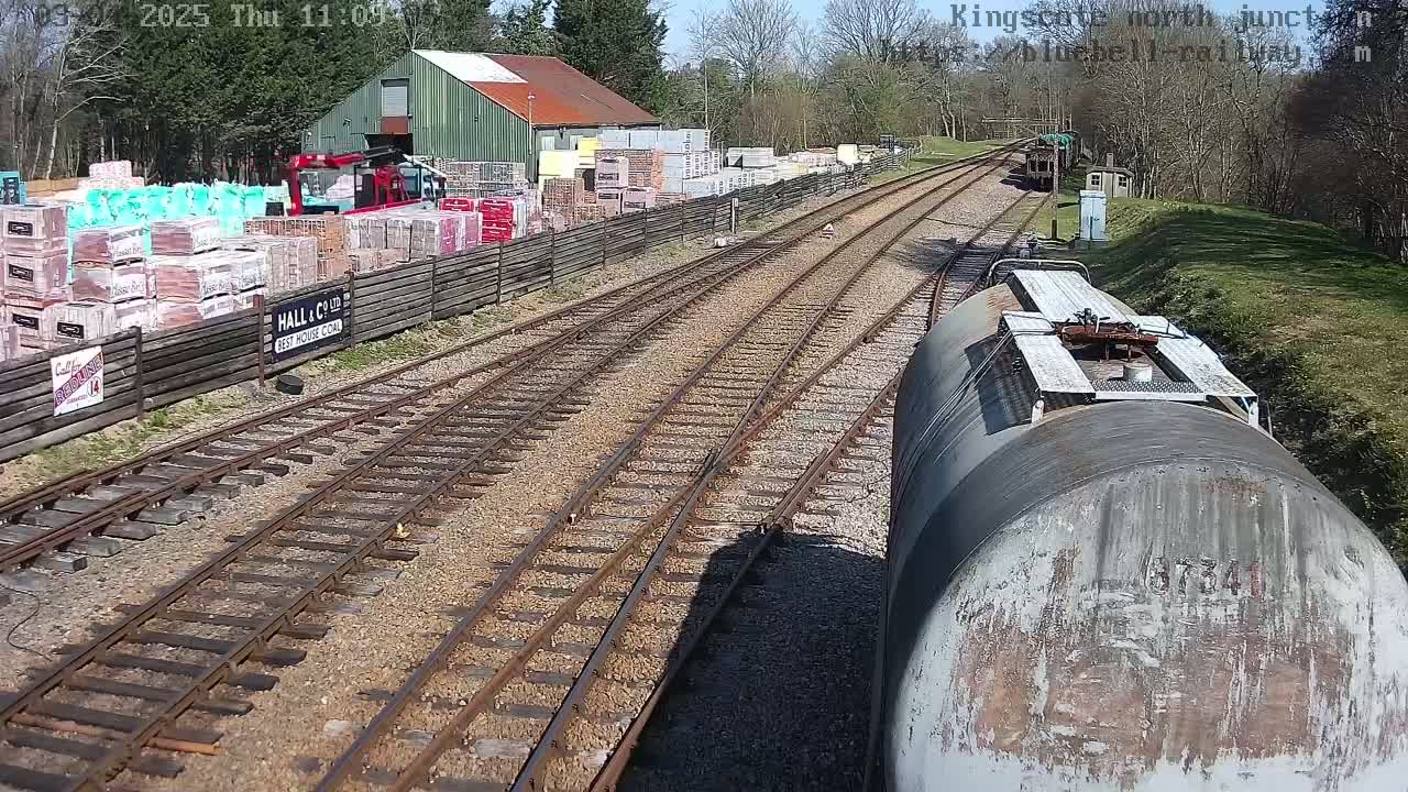 A train's perspective of railway tracks on a sunny day, passing a building and yard stocked with building materials.